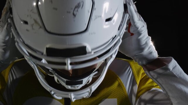 Close-up face portrait of young Caucasian male sportsman in yellow team jersey with shoulder pads putting on helmet for game of American football at dark arena, looking at camera with menacing stare