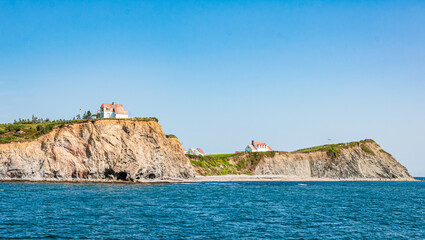 Rocky coastal cliffs with small houses above the sea in Gasp&eacute;sie near Perc&eacute;, Quebec, under a clear blue sky and calm ocean waters.
