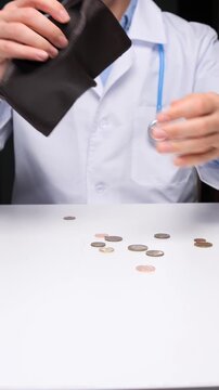 Vertical video doctor in white coat holds empty brown wallet as euro coins spill onto a white desk, symbolizing healthcare workers' low income, rising costs, debt and financial hardship