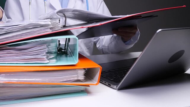 Doctor in a white coat managing a large stack of patient files and documents while using a laptop, representing the administrative workload and bureaucracy in healthcare