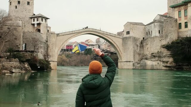 Woman from behind waving the flag of Bosnia and Herzegovina near the Stari Most bridge in Mostar. Travel lifestyle scene with river, historic architecture and national identity. Horizontal 4k footage