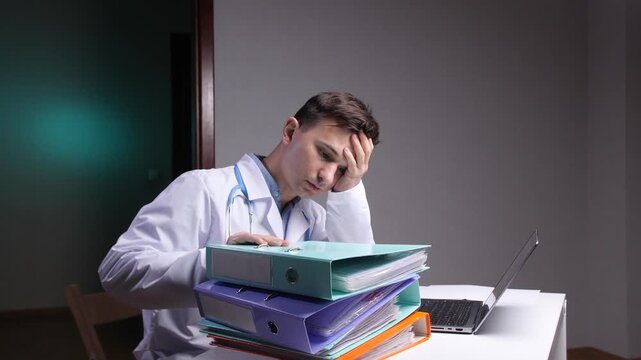 Overworked doctor wearing a lab coat and stethoscope experiencing burnout, resting his head on his hand while sitting at a desk late at night next to a stack of binders and a laptop
