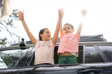 Joyful children standing through car sunroof with arms raised in nature. High quality photo