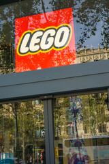 Fototapeta premium Barcelona, Spain, Europe - Nov 23, 2025: lego logo red yellow glass window storefront reflecting trees and historic architecture in a city street under blue sky with copy space for banner design