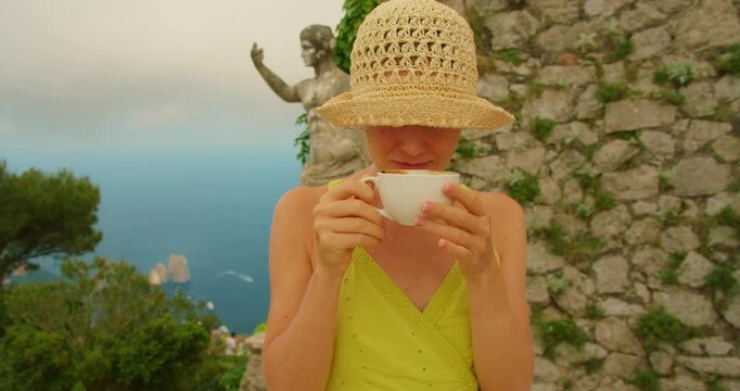 Woman enjoying a cup of coffee near statue of Augustus Caesar on the peak of Monte Solaro in Capri island with Faraglioni cliffs far below