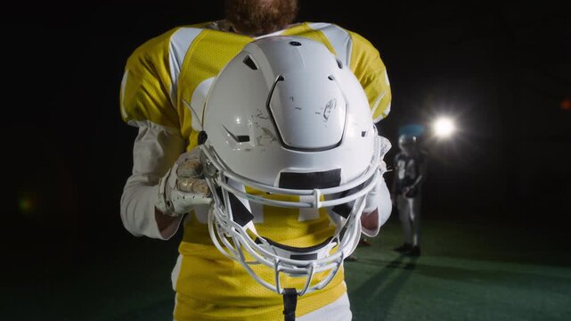 Medium midsection shot of anonymous male American football player, wearing yellow team jersey with chest and shoulder pads, posing at dark stadium, holding white helmet, before gridiron game