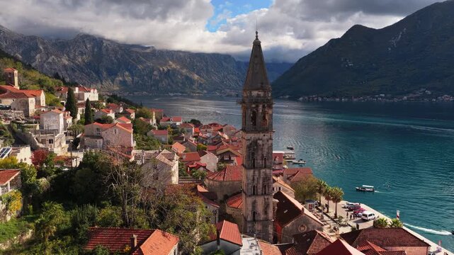 Aerial drone view of the bell tower in Perast rising above traditional stone houses, with the Bay of Kotor and steep mountains visible behind it. Bay of Kotor, Montenegro