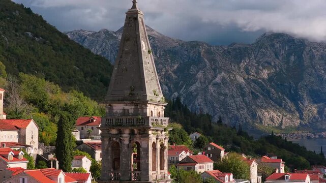 Aerial drone view of the bell tower in Perast rising above traditional stone houses, with the Bay of Kotor and steep mountains visible behind it. Bay of Kotor, Montenegro