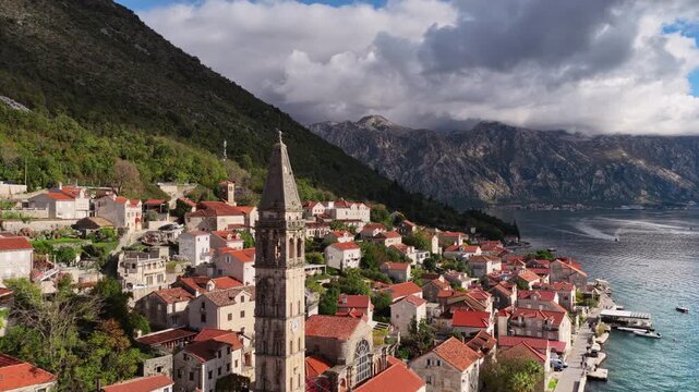 Aerial drone view of the bell tower in Perast rising above traditional stone houses, with the Bay of Kotor and steep mountains visible behind it. Bay of Kotor, Montenegro