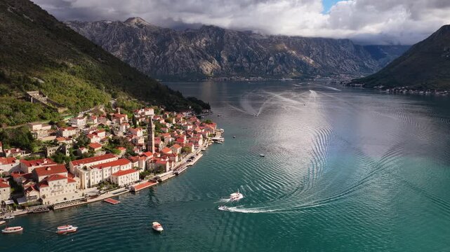 Aerial drone view of the hillside town of Perast, highlighting the tall church tower, dense red roofed houses, and the waterfront along the Bay of Kotor