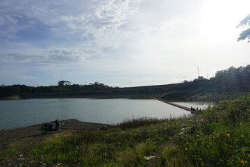 A lake with a clear sky above it. The water is calm and still. The trees surrounding the lake are lush and green, Malang, East Java, Indonesia.