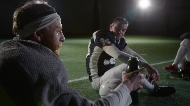 Medium shot of young Caucasian male gridiron football player passing bottle of water or energy drink to thirsty team mate during interval, while sitting together resting on turf