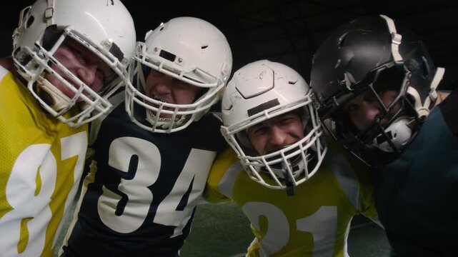 Medium portrait of four happy Caucasian male gridiron football team players standing together on pitch after game, wearing helmets and mouthguards, hugging, smiling and looking at camera