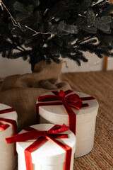 Christmas gifts wrapped in beige textured boxes with shiny red ribbons placed under a decorated Christmas tree with burlap skirt on woven rug in cozy home setting with warm natural daylight