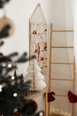 Advent calendar with numbered fabric pockets filled with candy canes in cozy minimalist room with wooden ladder red ribbons paper Christmas tree and fairy lights creating calm warm holiday atmosphere