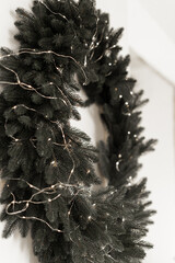 Close up of lush green Christmas wreath decorated with delicate string fairy lights on a white wall minimal winter holiday home decor with warm neutral tones and natural festive evergreen texture