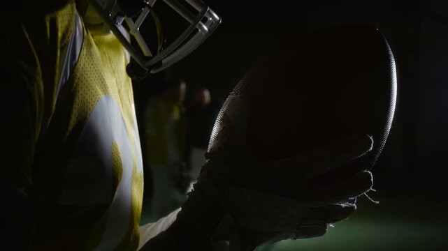 Side close-up of young Caucasian male quarterback preparing for game, touching face mask and helmet with ball, while getting in mood for training session with teammates