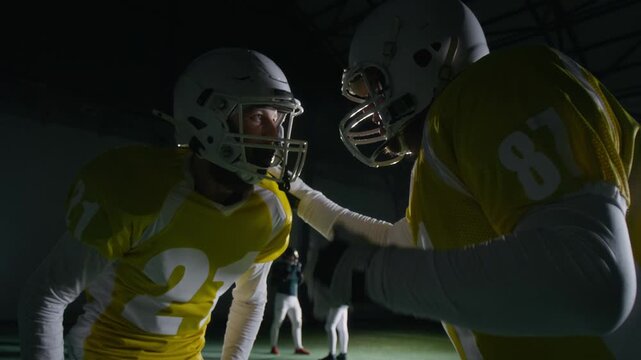 Medium shot of two male gridiron football players in helmets and padded body armor discussing attack or defense strategy, huddling then running off, during training game at dark sports arena