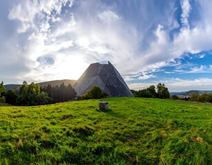 Pyramid-like structure on grassy hill under a bright sky with clouds and trees