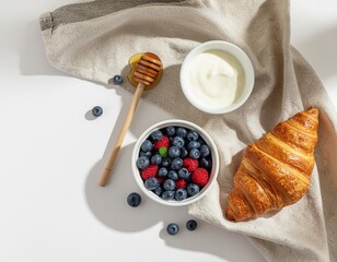 A delicious breakfast featuring fresh croissants and coffee served with a healthy bowl of yogurt, blueberries, and red berries on a white table with a glass of milk and a spoon