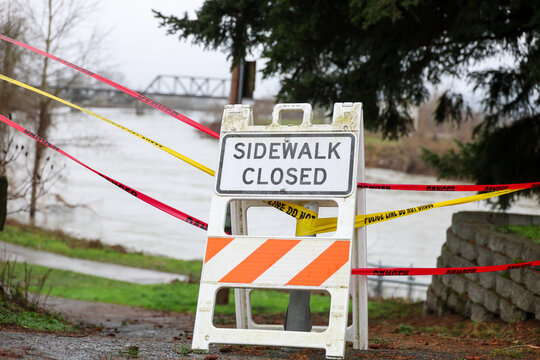 Sidewalk closed sign blocks foot traffic from a walking path above a deep river overflowing with flood water. The sign is for the safety of pedestrians during an emergency. Caution tape also attached.