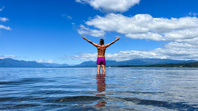 An unrecognizable sporty man stands in the fresh water of Lake Villarrica in southern Chile, holding his arms wide open above his head in a gesture of freedom and enjoyment. Mountains rise in the back