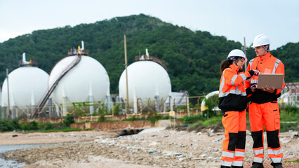 Two engineers in orange safety gear and hard hats stand on a coastline. They are using a laptop and...