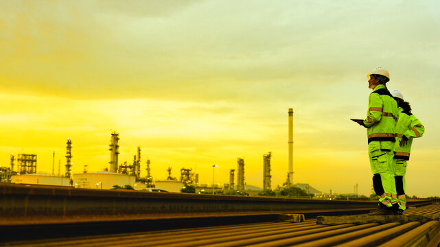 Two industrial workers in high visibility safety gear and hard hats stand on rusted metal pipes. They are inspecting a large factory or refinery plant during a golden sunset.