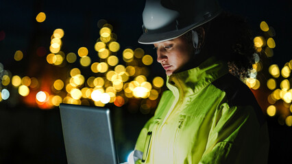 A close up of a female industrial worker in a hard hat and safety gear looking at a lit up factory or refinery at night. The background lights are blurred into bokeh.