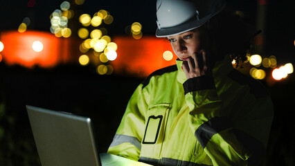 A petrochemical engineer in PPE monitors refinery operations during a night shift. The blurred...