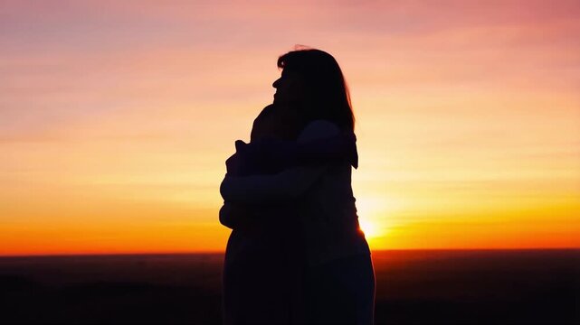 Silhouette of loving mother and child hugging tight against a dramatic orange sunset sky outdoors