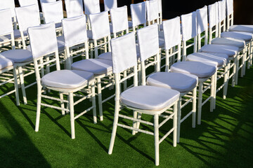 Outdoor rows of white Chiavari chairs with clean back covers on green lawn, sunlit and empty. Blank fabric ready for branding or text, ideal seating mockup for conference, wedding or event