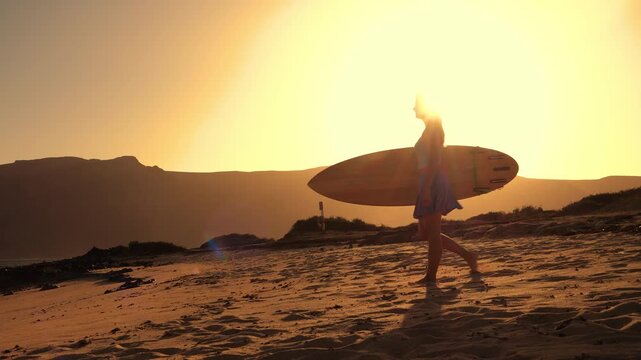 LENS FLARE, SILHOUETTE: Female surfer walks with her surfboard across a sandy beach at sunset. Woman wearing a skort goes for an evening surf session while on holidays at beautiful Lanzarote island.