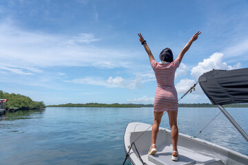 Woman watching a tropical sea and lagoon horizon from a boat bow, back view with arms raised in freedom pose, calm water, mangrove islands, blue sky and beach space, summer travel concept