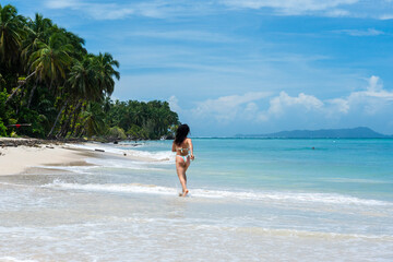 Obraz premium Woman jogging on tropical beach by turquoise ocean, back view in bikini with palm trees, wide seascape, blue sky, summer holiday travel lifestyle, tropical beach.