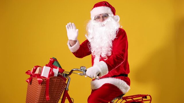 Santa Claus on bicycle with Christmas gifts in basket greeting someone against yellow background. Camera moving forward