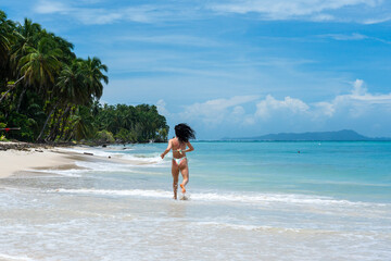 Obraz premium Woman jogging on tropical beach by turquoise ocean, back view in bikini with palm trees, wide seascape, blue sky, summer holiday travel lifestyle, tropical beach.