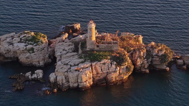 Aerial drone view of an isolated rocky Peristeres Island featuring the old lighthouse ruins, illuminated by golden hour sunlight against the deep blue Adriatic Sea. Greece