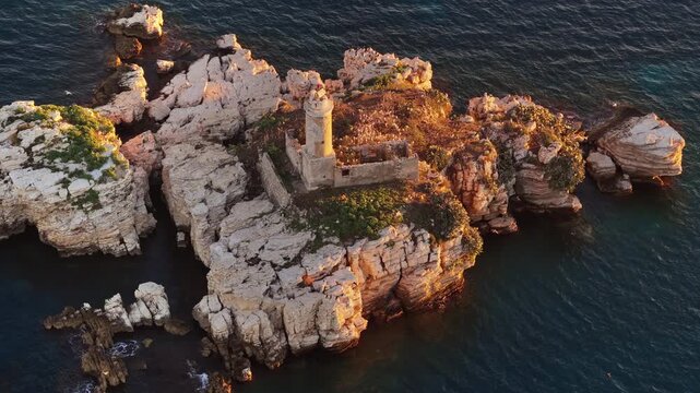 Aerial drone view of an isolated rocky Peristeres Island featuring the old lighthouse ruins, illuminated by golden hour sunlight against the deep blue Adriatic Sea. Greece