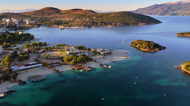 Aerial drone view over Ksamil town, capturing its buildings, beaches, clear waters, and lush hillsides illuminated by soft sunlight