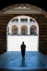 Unrecognized Latin woman walking the archway corridor of Museo Vivo del Muralismo in Mexico City, toward the courtyard adorned with murals and Mexican flags, surrounded by colonial stone architecture