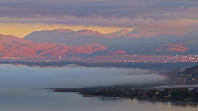 Aerial drone view of the Ohrid, Macedonia region, showing fog lifting from the wetlands and soft pink light falling on the surrounding mountains and villages