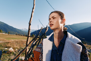 Woman portrait in mountains countryside outdoors wearing a warm jacket, sunlight on her face, young traveler near a wooden fence with scenic landscape and clear blue sky above. © SHOTPRIME STUDIO