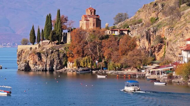 Aerial drone view of the Church of St. John at Kaneo, surrounded by rocky cliffs, autumn trees, and small boats docked along the shoreline