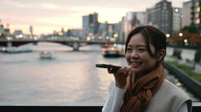 Smiling asian woman happily talking on her smartphone by the Sumida river in Tokyo at sunset