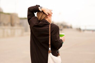 Woman in brown coat holding coffee cup outdoors on cloudy day. Natural feel with soft light. Casual, confident pose and expression add genuine and approachable vibe