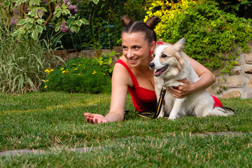 Cheerful woman smiles while holding happy white dog on green lawn in vibrant garden with plants and stone walls. Adoption from local dog shelters