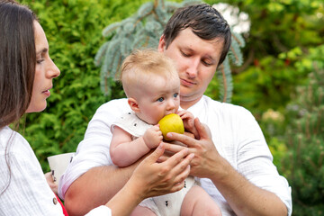 Close view of a baby gnawing a yellow apple while father supports and mother guides, an intimate feeding moment in a lush summer garden