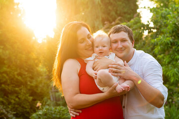 The couple leans in, hands supporting their grinning baby as the sun bursts behind them in a hazy flare
