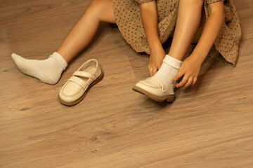 Young girl dressed in a polka dot dress sits on a wooden floor, carefully putting on beige shoes over white socks, highlighting a candid and everyday childhood moment indoors
